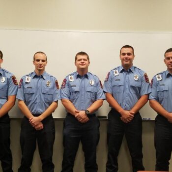 Five uniformed firefighters stand in a row in front of a whiteboard, wearing light blue shirts with badges and dark pants. They are posing for a group photo indoors.