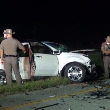 A nighttime scene shows a white SUV with front-end damage on the roadside. Three law enforcement officers in uniform examine the crash site, surrounded by debris, on a divided highway.
