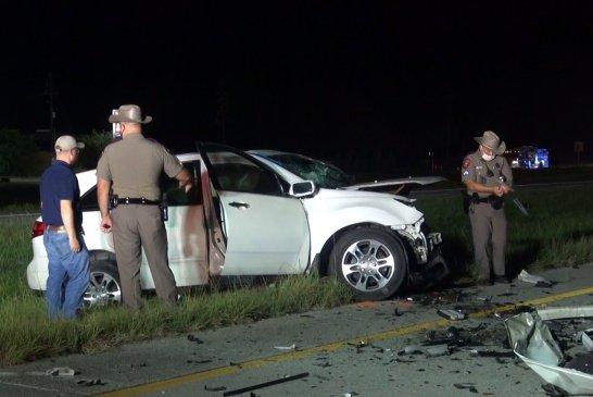 A nighttime scene shows a white SUV with front-end damage on the roadside. Three law enforcement officers in uniform examine the crash site, surrounded by debris, on a divided highway.