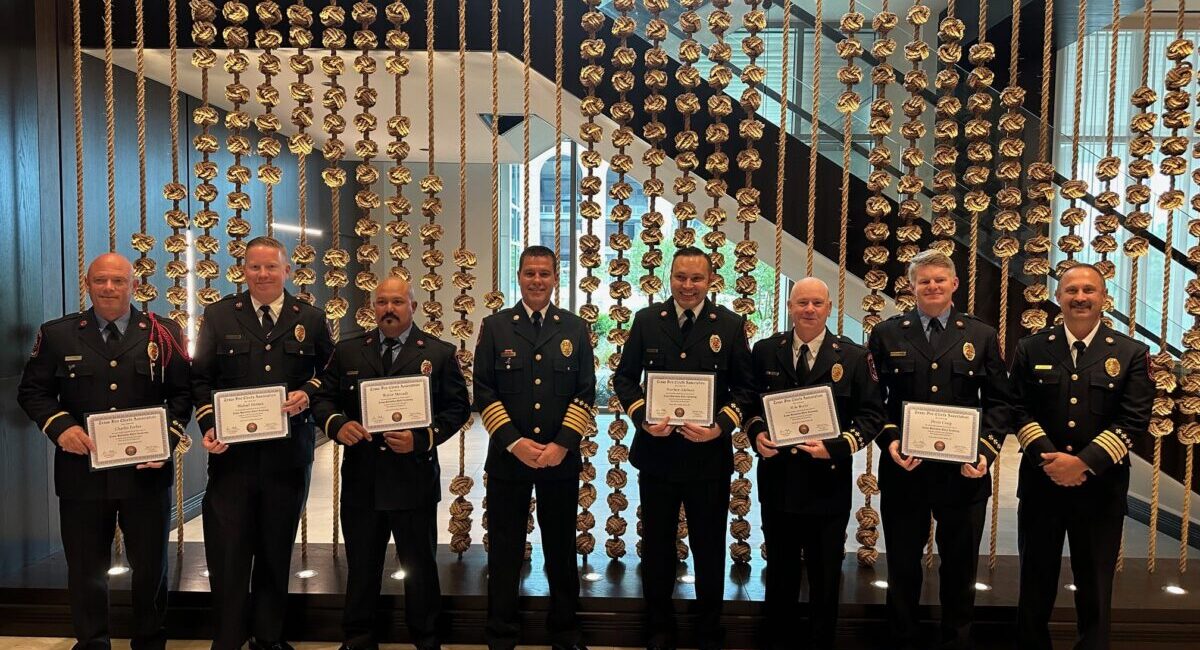 Eight uniformed firefighters stand in a line, each holding a certificate. They are in front of a decorative backdrop with chains and small, repetitive elements. A staircase is visible in the background.
