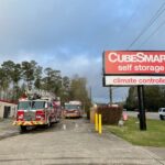 Fire trucks are parked outside a CubeSmart self-storage facility with a prominent sign that reads climate controlled. The background features trees and a cloudy sky.