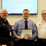 Three men stand indoors smiling. The man in the middle, wearing a blue uniform, holds a certificate and plaque. The other two men, wearing black and white uniforms, stand on either side of him. A screen displays text in the background.