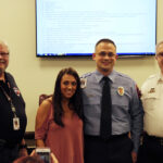 Four people stand indoors posing for a photo. A woman is flanked by two men in uniforms and one in a black shirt. A screen with text is visible in the background.
