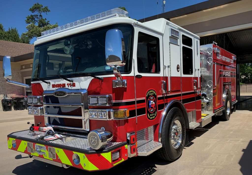 A red and white fire truck is parked outside a fire station. It has a shiny exterior with a prominent front grille and features black and white striping. The vehicle displays the text Engine 112 and has a detailed emblem on the side.
