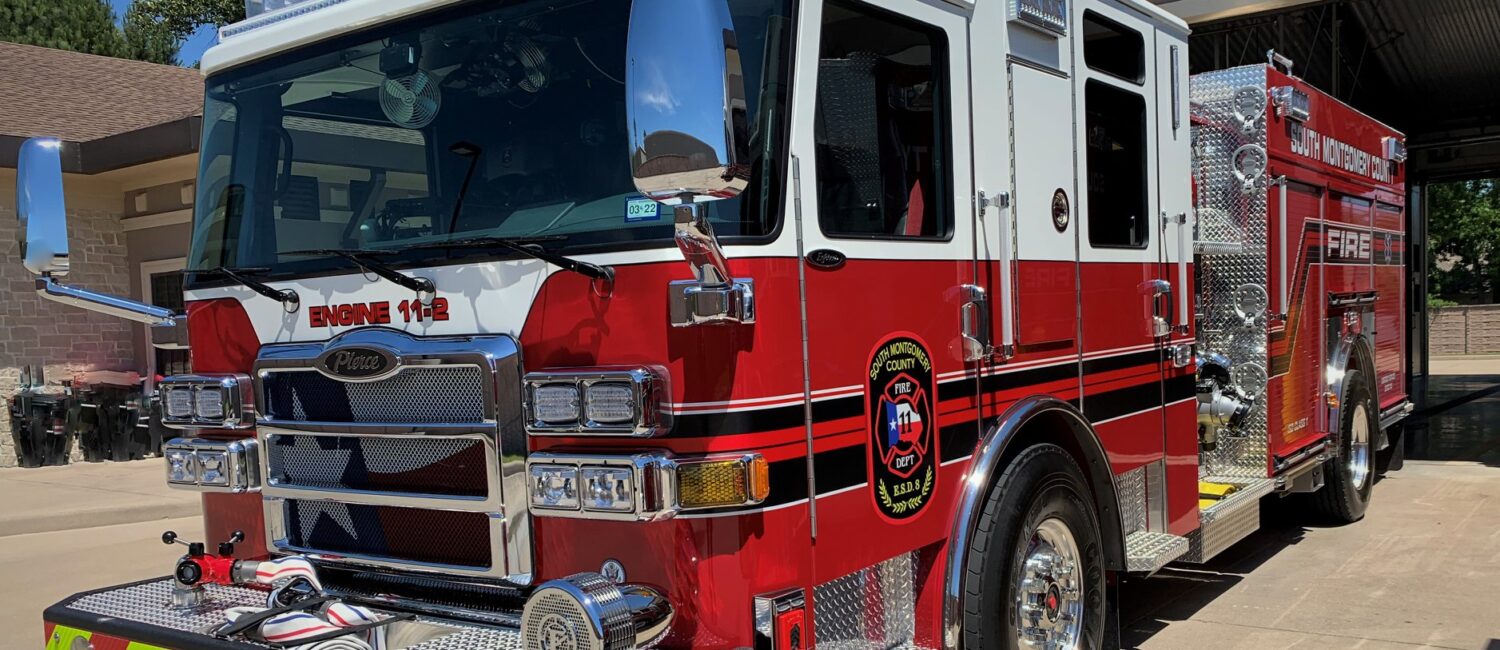 A red and white fire truck is parked outside a fire station. It has a shiny exterior with a prominent front grille and features black and white striping. The vehicle displays the text Engine 112 and has a detailed emblem on the side.