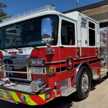 A red and white fire truck is parked outside a fire station. It has a shiny exterior with a prominent front grille and features black and white striping. The vehicle displays the text Engine 112 and has a detailed emblem on the side.