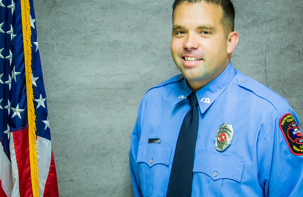 A person in a light blue uniform with a dark tie stands beside an American flag. The uniform has patches on the arms and a badge on the chest. The background is a textured gray.