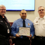 Three men stand together, smiling. The man in the center, wearing a blue uniform, holds a framed Certificate of Promotion award. The two other men wear white shirts with insignias. A presentation slide is visible in the background.