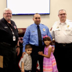 A firefighter in a blue uniform stands with two children and two men in white uniforms during a ceremony. The children, a boy and a girl, stand in the middle, the girl hugging the firefighter. An American and another flag are visible in the background.