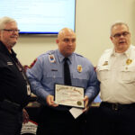 Three men stand together; the man in the center, wearing a blue uniform, holds a certificate. The two men on either side wear white shirts. A flag and other attendees are in the background.