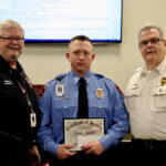 Three men stand indoors. The man in the middle, wearing a blue uniform, holds a Certificate of Service. The other two, in black and white uniforms, stand on either side of him. A presentation slide is visible in the background.