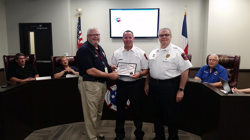 Three men stand in the center of a room, with one handing a framed certificate to another. Behind them, three people are seated at a conference table. Flags and a monitor are visible in the background.