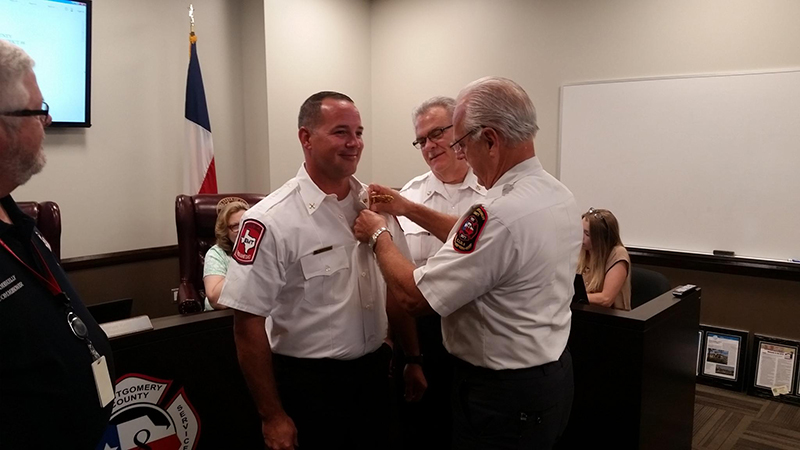 Three uniformed men stand in an office: one is pinned with a badge by another. A third man observes, smiling. A flag and seated people are in the background.
