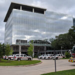 A modern glass office building is shown with several emergency vehicles, including fire trucks and SUVs, parked outside. Orange traffic cones line the driveway, and trees surround the scene under a cloudy sky.