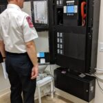A man in a uniform stands next to an open fire alarm control panel in a room with visible wiring and pipes. He appears to be inspecting or operating the equipment.