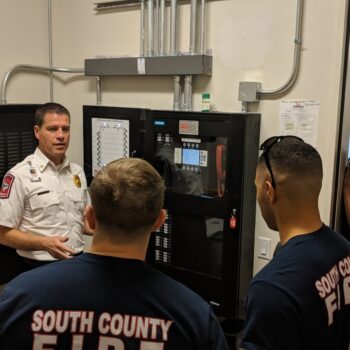 A group of firefighters gathers in a room with electronic equipment. A person in a white uniform is instructing the group, who are wearing shirts reading South County Fire. They appear to be discussing the control panel in the room.