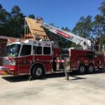 A fire truck with ladder L114 is extended to the roof of a building under construction. Firefighters on the roof and ladder are working under a clear blue sky, surrounded by trees.