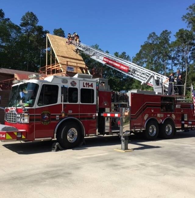 A fire truck with ladder L114 is extended to the roof of a building under construction. Firefighters on the roof and ladder are working under a clear blue sky, surrounded by trees.