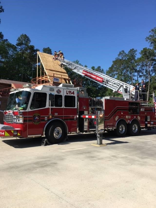 A fire truck with ladder L114 is extended to the roof of a building under construction. Firefighters on the roof and ladder are working under a clear blue sky, surrounded by trees.