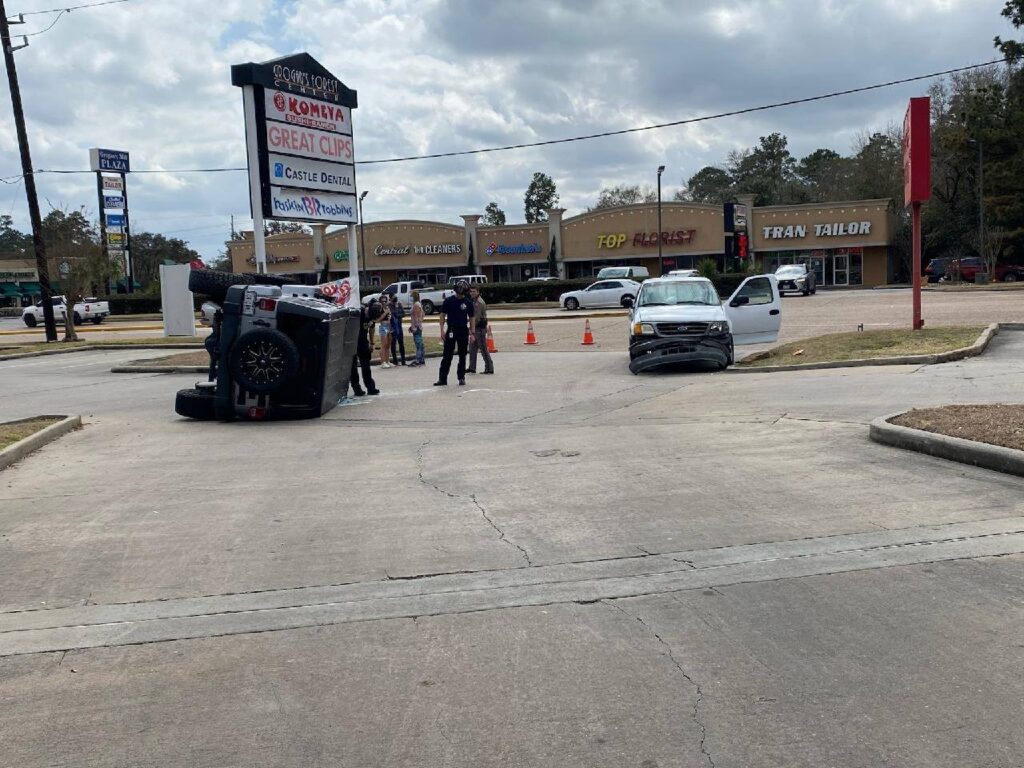 A car is flipped on its side in a parking lot, with people standing nearby. A second car is parked close, facing the flipped vehicle. Several businesses, such as a nail salon and tailor, are visible in the background.