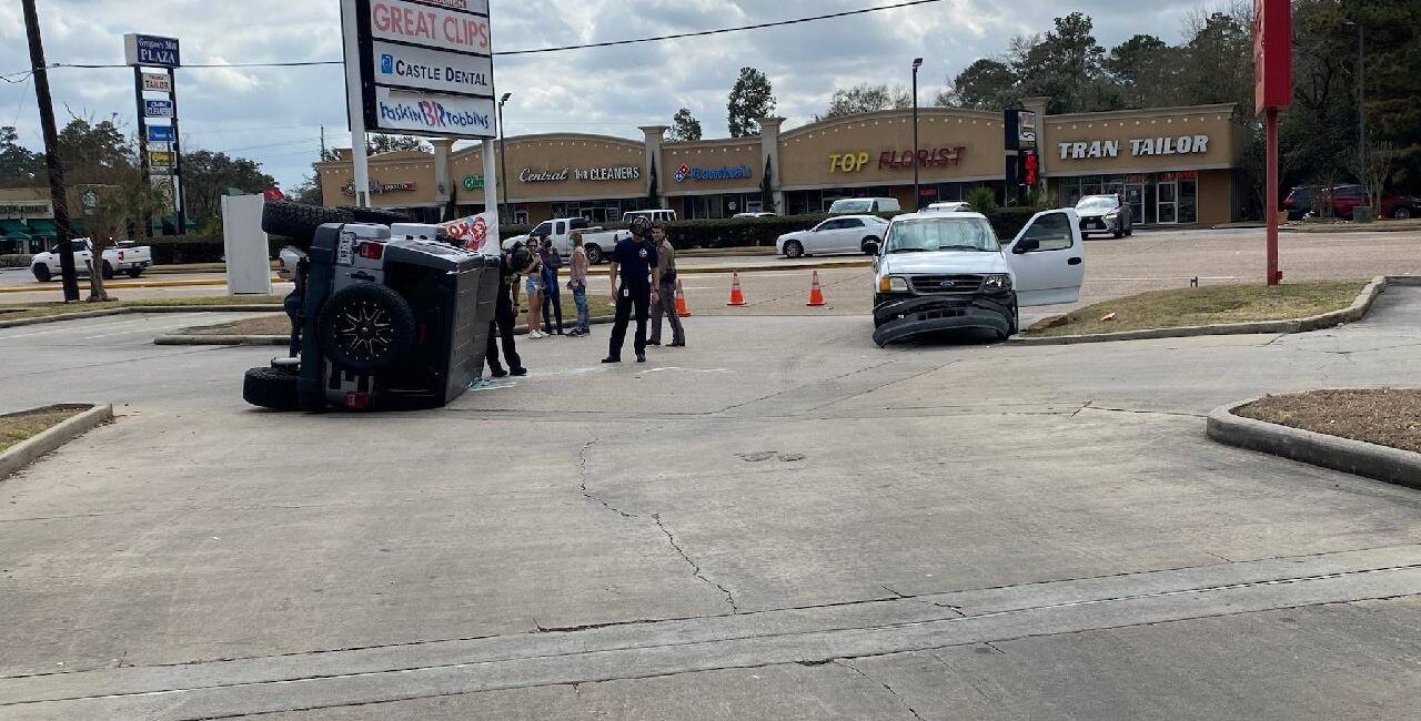 A car is flipped on its side in a parking lot, with people standing nearby. A second car is parked close, facing the flipped vehicle. Several businesses, such as a nail salon and tailor, are visible in the background.