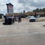 A car is flipped on its side in a parking lot, with people standing nearby. A second car is parked close, facing the flipped vehicle. Several businesses, such as a nail salon and tailor, are visible in the background.