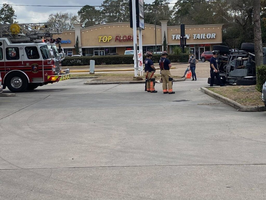 A fire truck and several firefighters stand near an overturned vehicle on a concrete surface. Nearby, a few people observe the scene. In the background, there are shops, including Top Floor and Tran Tailor.
