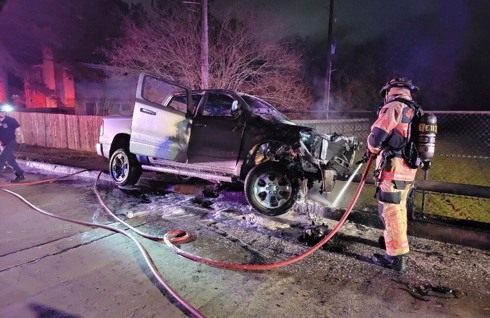 A firefighter sprays water on a white pickup truck with severe front damage, parked on a roadside at night. Hoses are on the ground, with emergency lights illuminating the scene. A fence and leafless tree are in the background.
