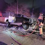 A firefighter sprays water on a white pickup truck with severe front damage, parked on a roadside at night. Hoses are on the ground, with emergency lights illuminating the scene. A fence and leafless tree are in the background.