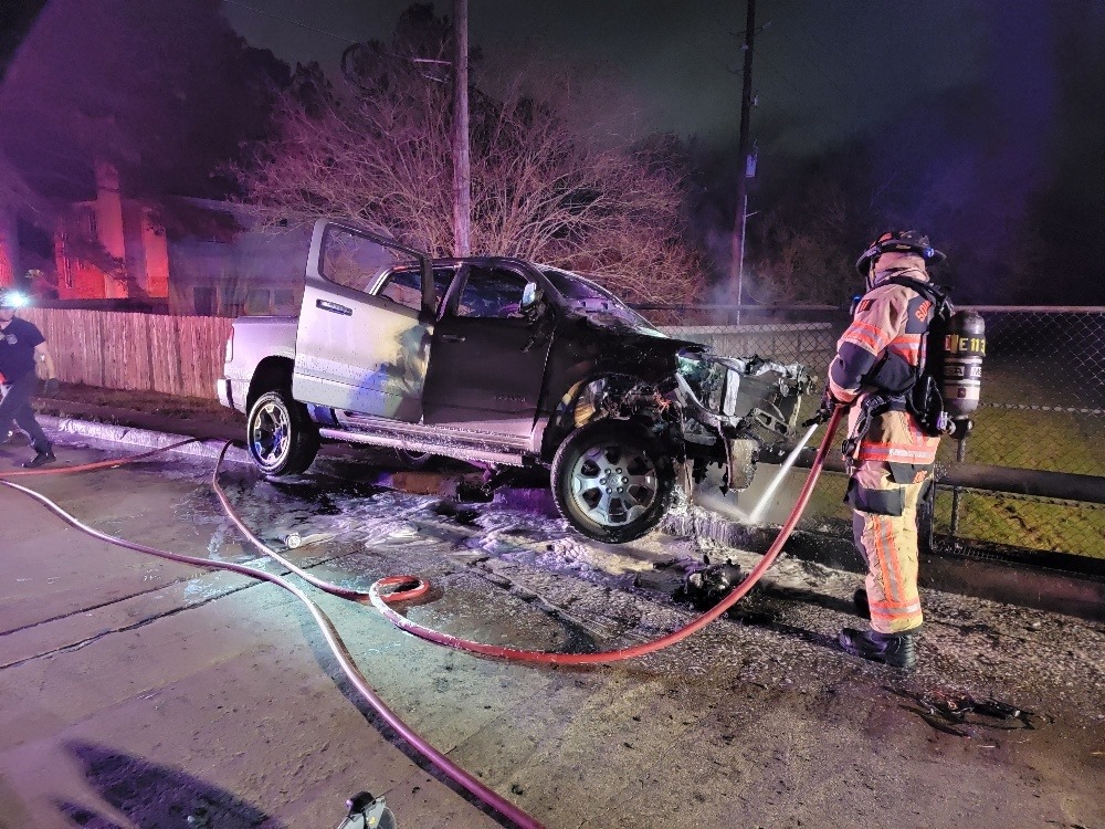 A firefighter sprays water on a white pickup truck with severe front damage, parked on a roadside at night. Hoses are on the ground, with emergency lights illuminating the scene. A fence and leafless tree are in the background.