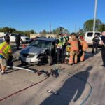 A car accident scene on a highway involving a silver car. Emergency responders, including firefighters and police officers, are at work. A FedEx truck and another vehicle are parked nearby. Debris is scattered on the road under clear blue skies.