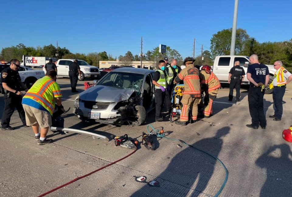 A car accident scene on a highway involving a silver car. Emergency responders, including firefighters and police officers, are at work. A FedEx truck and another vehicle are parked nearby. Debris is scattered on the road under clear blue skies.