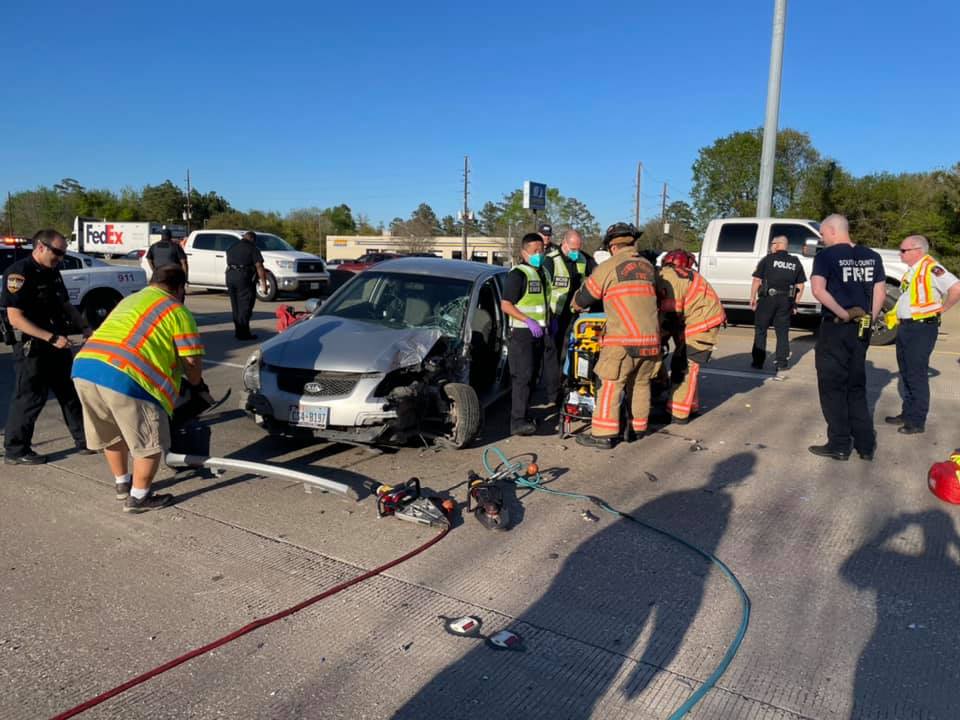 A car accident scene on a highway involving a silver car. Emergency responders, including firefighters and police officers, are at work. A FedEx truck and another vehicle are parked nearby. Debris is scattered on the road under clear blue skies.