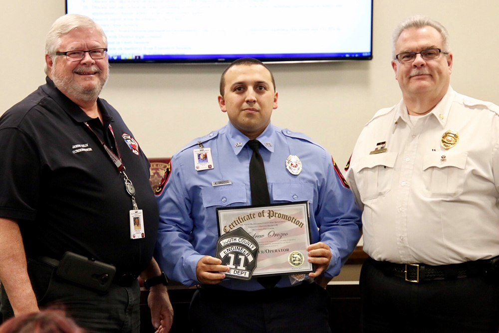 Three men stand together in a room. The man in the middle, wearing a blue uniform, holds a Certificate of Promotion. The two other men wear black and white uniforms, respectively, and smile at the camera. A screen and people are visible in the background.
