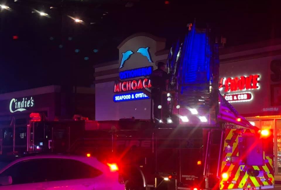 A fire truck with flashing lights is parked in front of a strip mall at night. Signs for Ostioneria Michoacan and Cindies are visible. A person stands atop the truck’s ladder, which is extended toward the building.