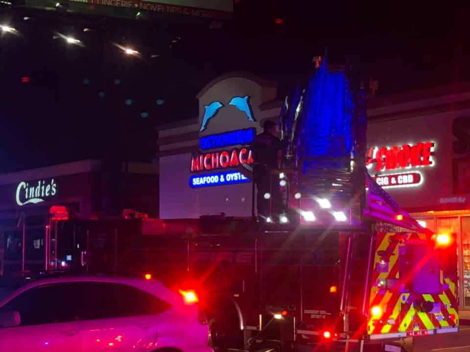 A fire truck with flashing lights is parked in front of a strip mall at night. Signs for Ostioneria Michoacan and Cindies are visible. A person stands atop the truck’s ladder, which is extended toward the building.