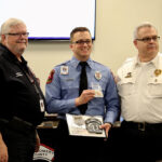 Three men in uniform stand in a room with a screen and flag in the background. The man in the center, holding a badge and award, smiles widely. The others flank him, also smiling, wearing fire department uniforms.