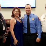 Four people are posing indoors, including two men in uniform, one in a blue shirt and the other in white. Theyre standing together smiling, with a woman in a blue dress among them. A presentation screen is visible in the background.