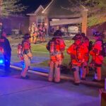 Firefighters in full gear stand by a house at night, with some carrying hoses towards the entrance. A fire truck and emergency vehicle are visible, and the scene is lit by red and blue lights. The garage door is open, revealing a parked car.