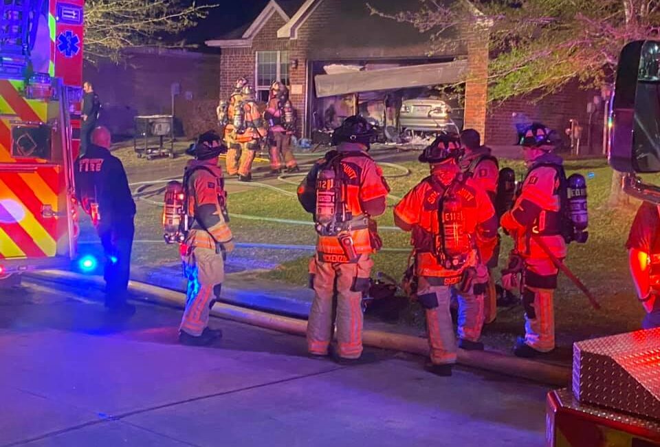 Firefighters in full gear stand by a house at night, with some carrying hoses towards the entrance. A fire truck and emergency vehicle are visible, and the scene is lit by red and blue lights. The garage door is open, revealing a parked car.