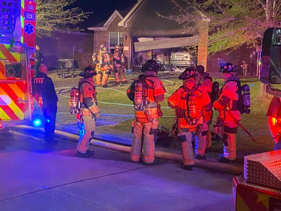 Firefighters in full gear stand by a house at night, with some carrying hoses towards the entrance. A fire truck and emergency vehicle are visible, and the scene is lit by red and blue lights. The garage door is open, revealing a parked car.