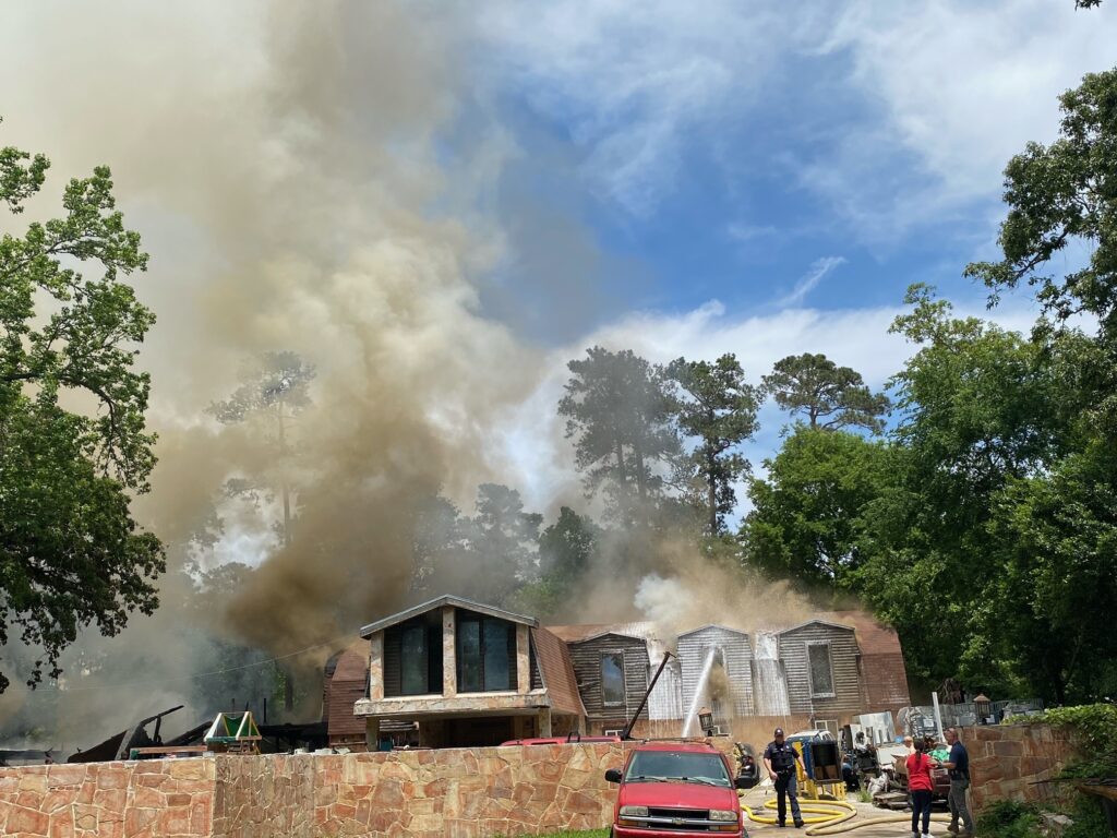 A house with wooden siding is engulfed in thick smoke, partially obscuring the blue sky. Firefighters are actively working to control the blaze. A red truck and a few people are in the foreground, with trees surrounding the scene.