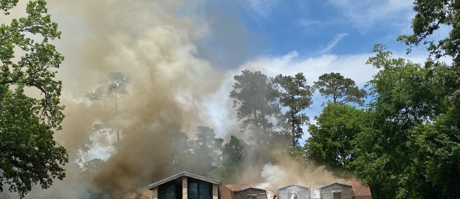 A house with wooden siding is engulfed in thick smoke, partially obscuring the blue sky. Firefighters are actively working to control the blaze. A red truck and a few people are in the foreground, with trees surrounding the scene.