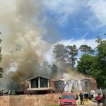 A house with wooden siding is engulfed in thick smoke, partially obscuring the blue sky. Firefighters are actively working to control the blaze. A red truck and a few people are in the foreground, with trees surrounding the scene.