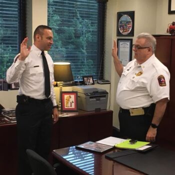 Two uniformed men are standing in an office, raising their right hands as if taking an oath. One is behind a desk with a green clipboard, while the other stands in front. The room has windows, and shelves with various items and decorations.