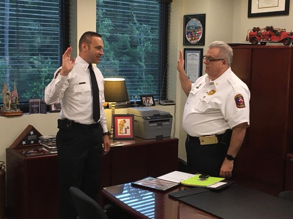Two uniformed men are standing in an office, raising their right hands as if taking an oath. One is behind a desk with a green clipboard, while the other stands in front. The room has windows, and shelves with various items and decorations.
