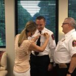 A woman is pinning a badge on the uniform of a man in the center, while another man in a uniform smiles beside them. They are in an office setting with large windows showing greenery outside.