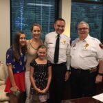 A family poses with two uniformed fire officers in an office. A woman and two girls stand next to the officers, all smiling. The office has a large window and framed certificates on the walls.