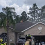 Firefighters, dressed in protective gear, are working on the roof of a house with smoke rising around them. A few other firefighters are gathered near the garage area of the single-story home, surrounded by trees under a cloudy sky.
