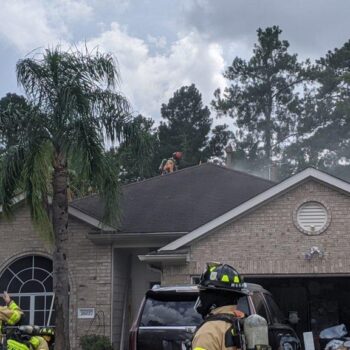 Firefighters, dressed in protective gear, are working on the roof of a house with smoke rising around them. A few other firefighters are gathered near the garage area of the single-story home, surrounded by trees under a cloudy sky.